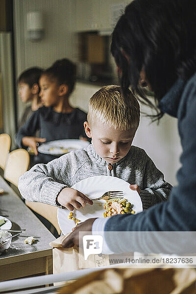 Männlicher Schüler wirft Essensreste in eine Papiertüte  die von einer Lehrerin in der Kindertagesstätte gehalten wird