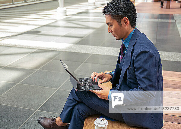 A young businessman in a blue suit on the move in a city downtown area  sitting on a bench using a digital tablet.