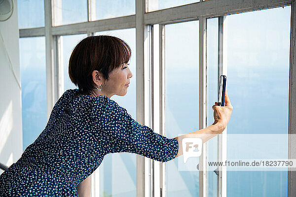 A mature Japanese woman using her mobile phone to take pictures from a viewing platform of the city and landscape below.