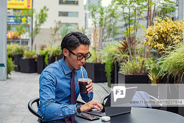 A young businessman in the city  on the move  a man seated at a cafe table outdoors  using a laptop  sipping a soft drink with a straw.