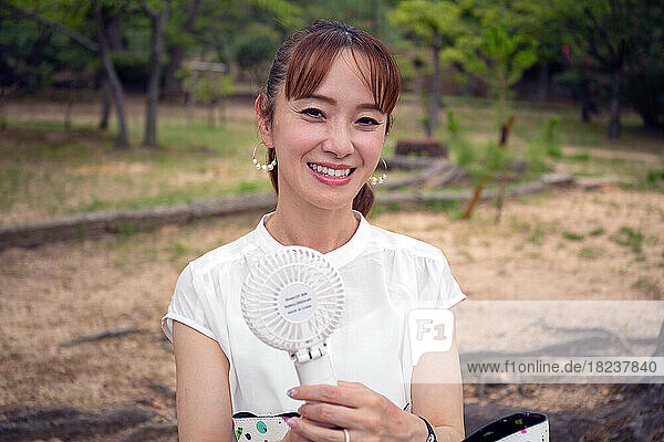 A mature Japanese woman outdoors in a park on a hot day holding a small electric fan.
