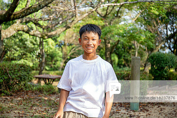 A 13 year old boy in a white teeshirt  smiling  outdoors in a park.