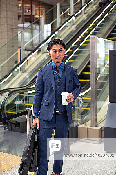 A young businessman in a blue suit on the move in a city downtown area  carrying a briefcase and cup of coffee.