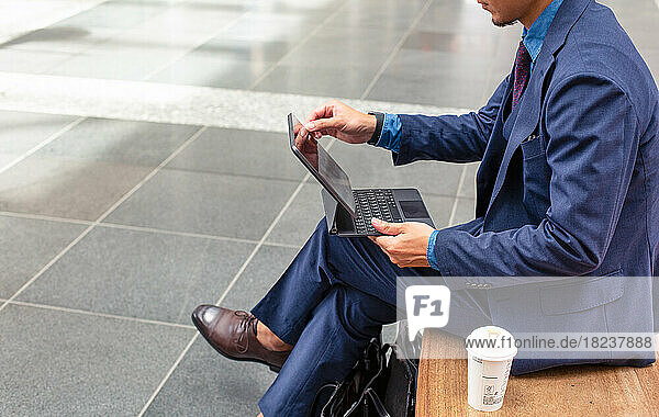 A young businessman in a blue suit on the move in a city downtown area  sitting on a bench using a digital touch screen tablet.
