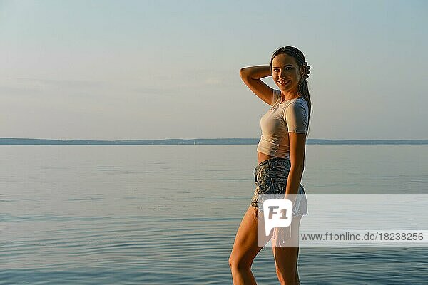 Glückliche junge Frau in weißem Tanktop und Jeansshorts am Strand bei Sonnenuntergang