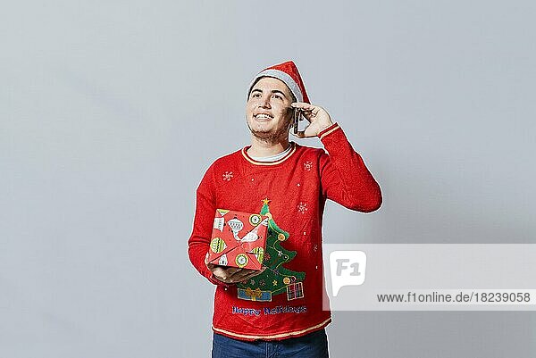 Happy guy holding gift and calling family at christmas  Smiling guy in christmas hat holding christmas gift box and talking on the phone  Christmas man holding gift and calling family on phone