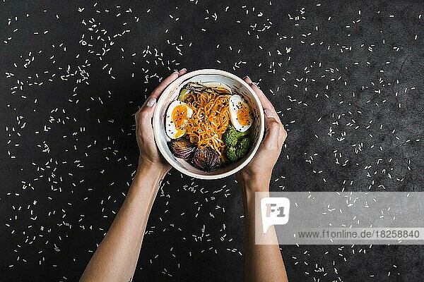 Erhöhte Ansicht Hände halten Schalen Ramen-Nudeln mit Eiern Salat Ausbreitung mit Reiskörnern schwarzen Hintergrund