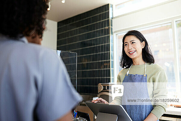 Smiling cafe owner with apron talking to customer at checkout counter