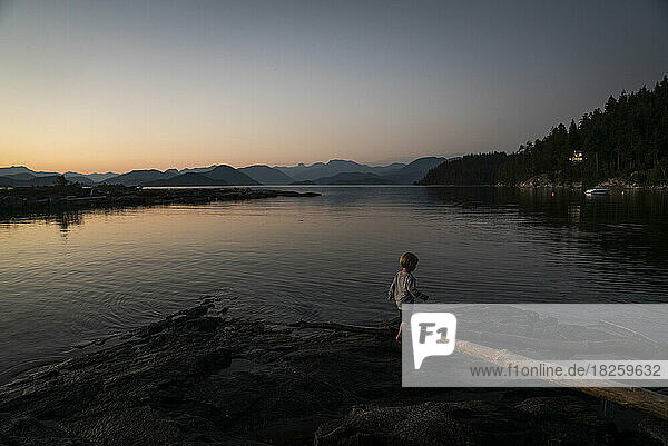 A boy walks on rocks at waters edge at dusk with mountains in back
