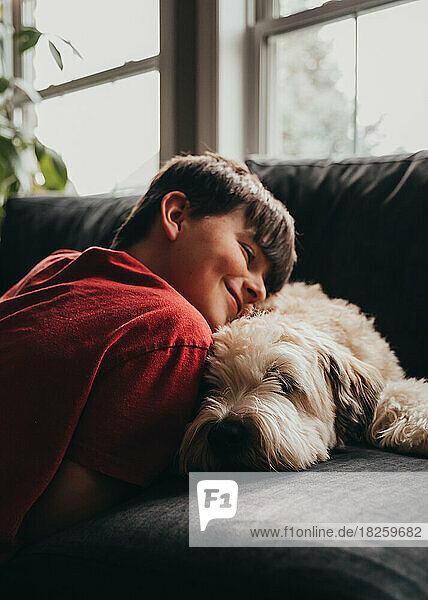 Happy smiling boy snuggling with fluffy dog on a sofa at home.