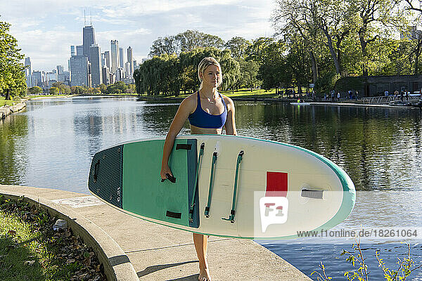 Woman carrying paddleboard walking at riverbank on sunny day