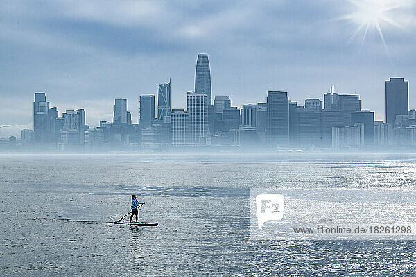 Woman paddleboarding in bay while city skyline in background