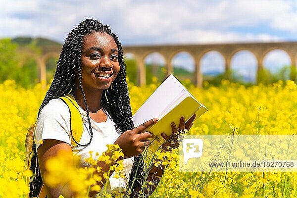 Ein Buch in der Natur lesend  lächelnd  ein schwarzes ethnisches Mädchen mit Zöpfen  eine Reisende  in einem Feld mit gelben Blumen