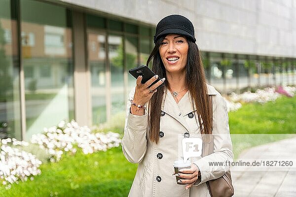 Erwachsene Geschäftsfrau vor dem Büro mit einem Kaffee in der Hand  die mit dem Telefon eine Sprachnachricht sendet