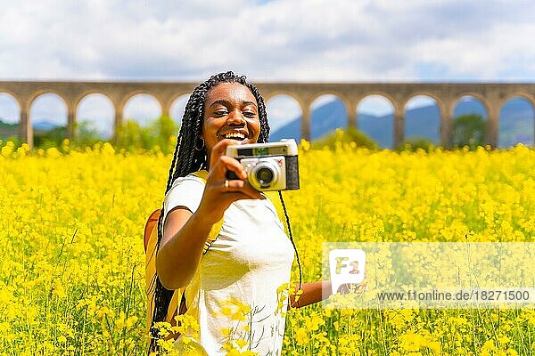 Mit einer alten Kamera lächelnd fotografieren  ein schwarzes ethnisches Mädchen mit Zöpfen  eine Reisende  in einem Feld mit gelben Blumen