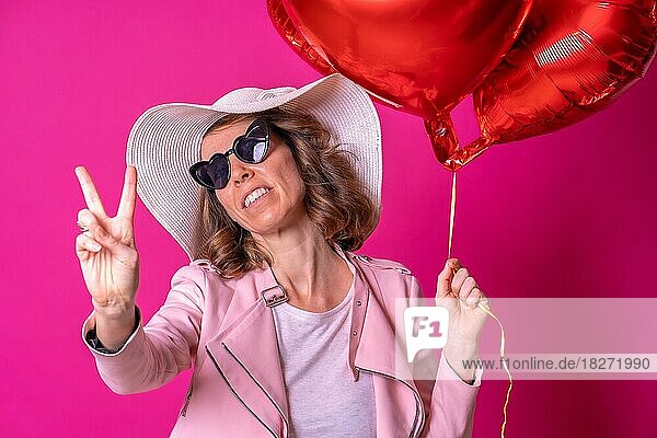 Blonde kaukasische Frau mit weißem Hut und Sonnenbrille macht das Siegeszeichen mit ihren Händen in einem Nachtclub mit einigen Herzluftballons  die ein Selfie machen  rosa Hintergrund