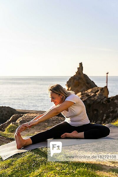 Eine blonde Frau macht Yoga-Übungen in der Natur am Meer und dehnt sich neben einem Leuchtturm