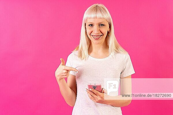 Blondes kaukasisches Mädchen im Studio auf rosa Hintergrund  Kopierraum  lächelnd auf das Handy zeigend