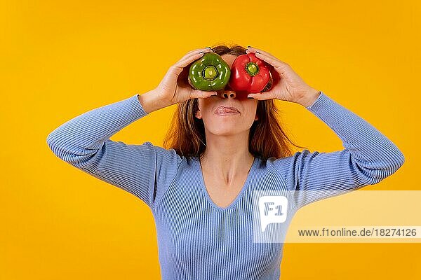 Vegetarische Frau im Porträt mit Paprika in den Augen auf gelbem Hintergrund  die ihre Zunge herausstreckt