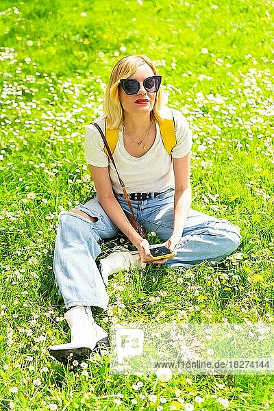 Porträt einer blonden Touristin mit Hut und Sonnenbrille  die im Frühjahr im Gras neben Gänseblümchen sitzt  in die Kamera schaut und die Natur genießt