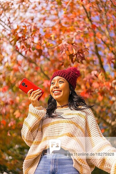 Asiatisches Mädchen im Herbst mit einem Handy  das lächelnd eine Sprachnachricht in einem Wald aus roten Blättern sendet  Technik