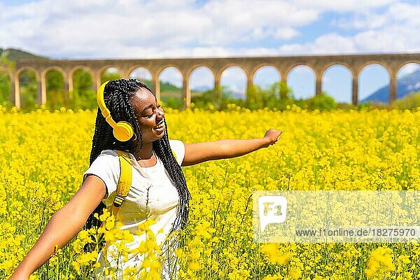 Musik hörend mit gelben Kopfhörern und geschlossenen Augen  ein schwarzes ethnisches Mädchen mit Zöpfen  eine Reisende  in einem Feld mit gelben Blumen