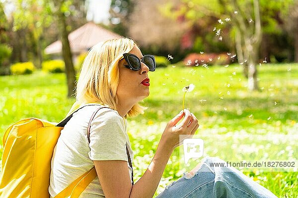 A tourist blonde woman blowing dandelion plant  wearing a hat and sunglasses sitting on the grass in spring next to daisies in a park in the city