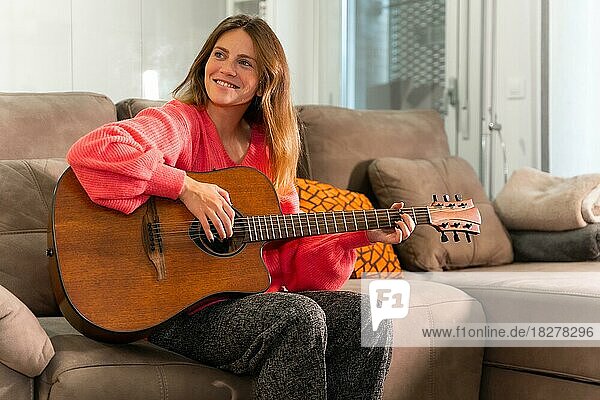 Frau spielt zu Hause Gitarre und sitzt lächelnd auf dem Sofa in ihrem Wohnzimmer neben dem Verkauf