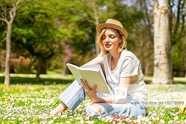 Ein junges blondes Mädchen mit Hut liest im Frühling in einem Park in der Stadt ein Buch  macht Urlaub in der Natur und neben Gänseblümchen  sitzt im Gras