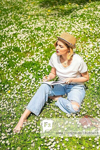 Ein junges blondes Mädchen mit Hut  das im Frühling in einem Park in der Stadt  in der Natur  auf dem Gras neben Gänseblümchen sitzend  Gedichte in ein Notizbuch schreibt