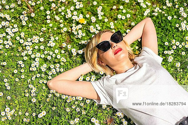 Ein junges blondes Mädchen mit Hut und Sonnenbrille atmet im Frühling in einem Park in der Stadt  Natur  auf dem Gras liegend neben Gänseblümchen  Teil Kopie Raum