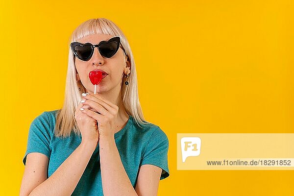 Mit Spaß essen ein Herz Lutscher  blonde kaukasische Mädchen auf gelbem Hintergrund Studio