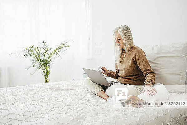 Reife Frau benutzt Laptop und streichelt Hund zu Hause im Bett