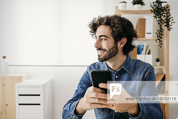 Thoughtful smiling young engineer holding smart phone sitting at desk in office