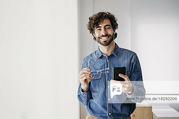 Happy young entrepreneur with smart phone standing by window in office
