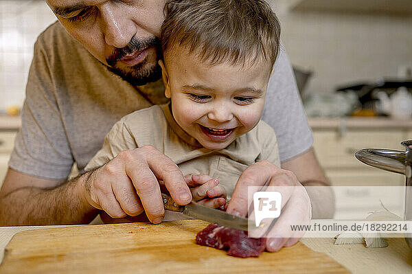 Father cutting red meat with son in kitchen