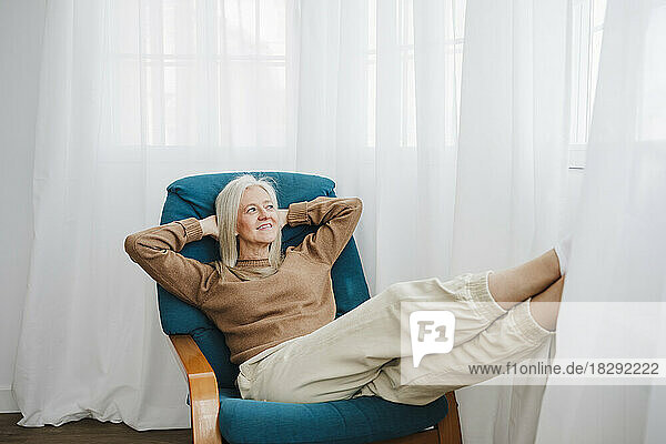 Smiling mature woman with hands behind head relaxing on armchair