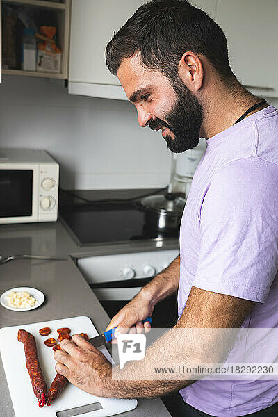 Lächelnder Mann schneidet Chorizo-Wurst an der Küchentheke