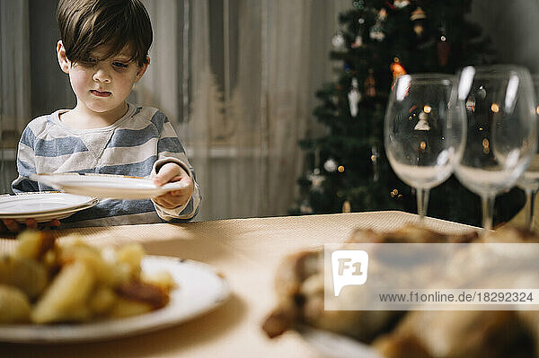 Cute boy arranging plates on dining table at home