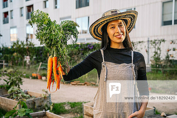 Happy woman holding carrots standing in urban garden