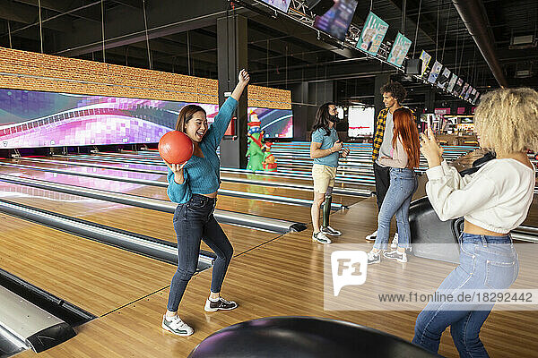Young woman taking photo of friend holding bowling ball at alley