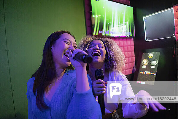 Happy young friends singing karaoke in microphone at amusement arcade