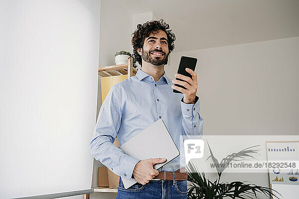 Contemplative young engineer holding mobile phone and laptop in office