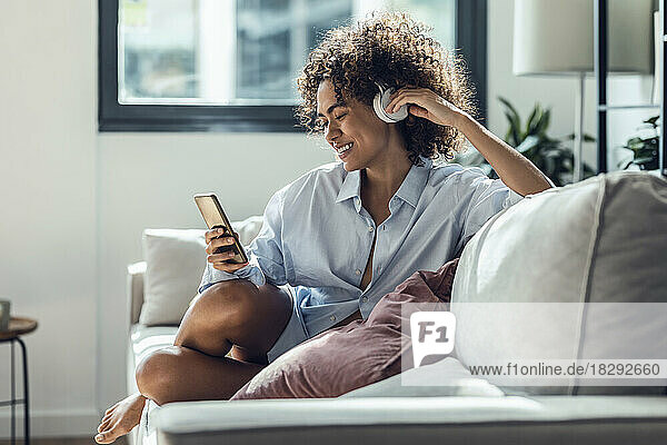 Smiling woman with headphones using smart phone on sofa at home