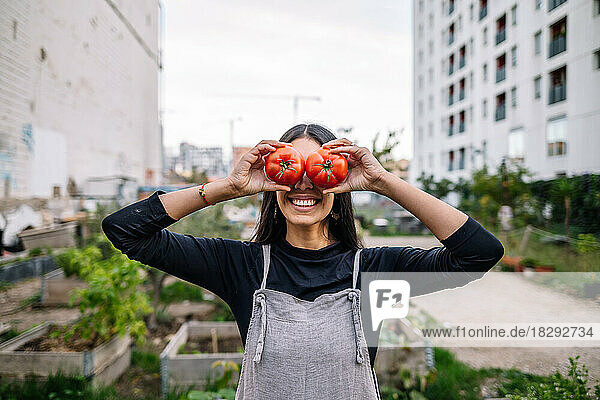 Happy woman holding tomatoes over eyes in urban garden