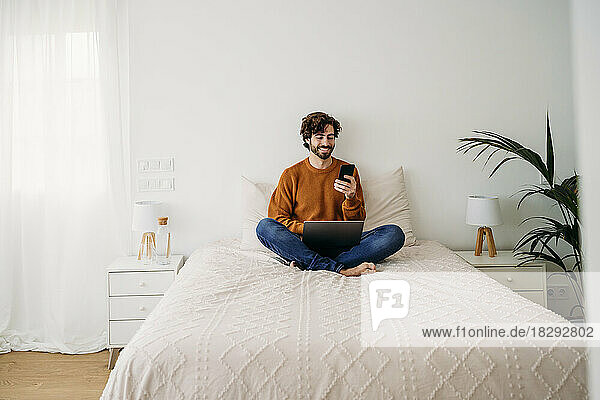 Happy freelancer using smart phone on bed at home