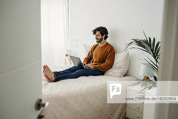 Smiling freelancer sitting on bed and working on laptop