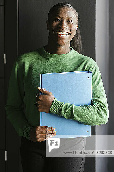 Cheerful businesswoman holding file folder standing in front of wall