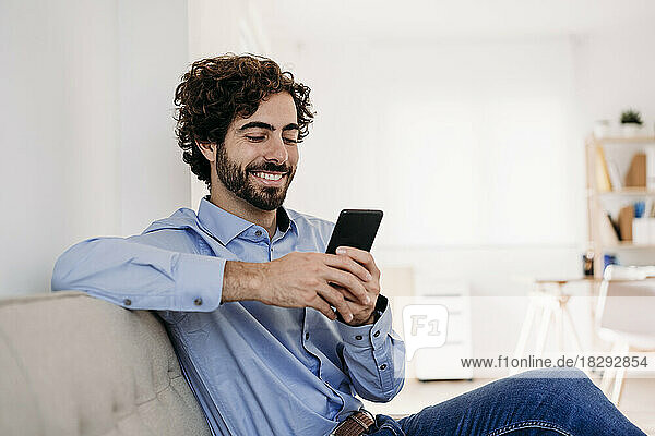 Happy young businessman using smart phone sitting on sofa in office