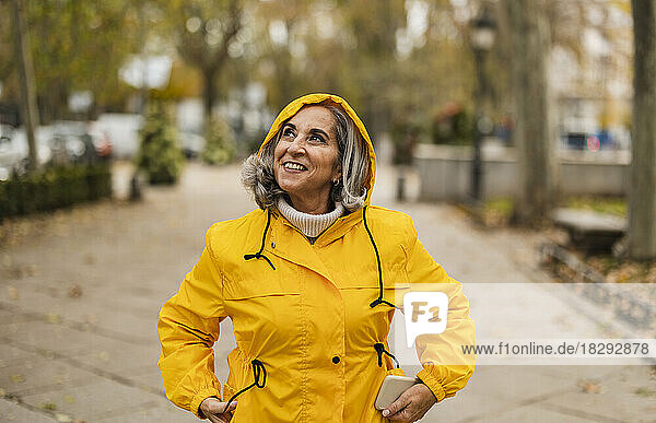 Nachdenkliche ältere Frau im gelben Regenmantel steht am Fußweg